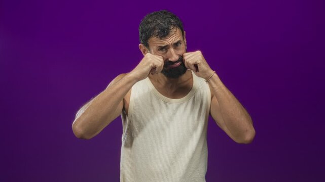 Man middle age bearded rubs eyes with hands and bare forearms visible in studio purple set; weariness.