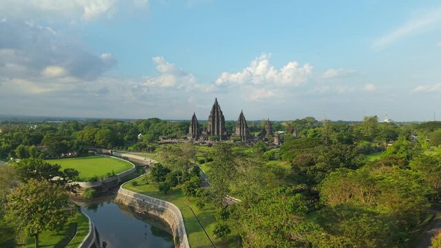 Aerial view of Prambanan Temple surrounded by lush green trees and a water canal under a cloudy sky in Kecamatan Prambanan, Daerah Istimewa Yogya, Indonesia.