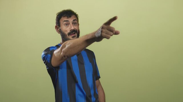 Man soccer player in blue striped uniform points finger forward in studio, open mouth and assertive stance; determination focus command.