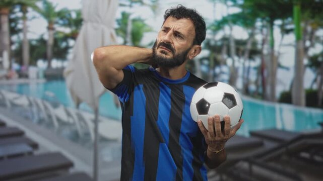 Man holding soccer ball with hand on neck and grimace at building poolside near lounge chairs and umbrella; injury pain recovery.