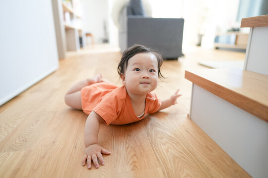 Tiny child crawls on warm wooden floor. Eyes bright, curious, unbothered by surroundings. Soft orange outfit adds cheerful contrast. Natural light streams through nearby window