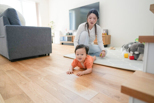 Mother smiles warmly while watching her baby crawl. Child wears bright orange shirt, focused on moving forward. Soft light fills cozy living room with wooden floors