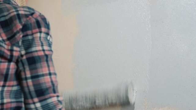A person painting a wall with a roller, applying light grey paint to create a fresh and smooth surface in a room.