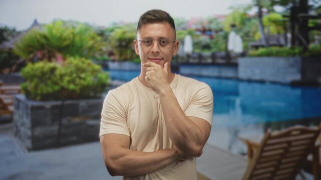 Man with glasses touching chin with hand in pensive pose by poolside lounge chair at building wearing beige t shirt; contemplation decision.