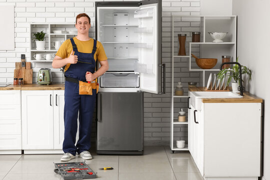 Happy male worker of repair service near fridge showing thumb-up gesture in kitchen