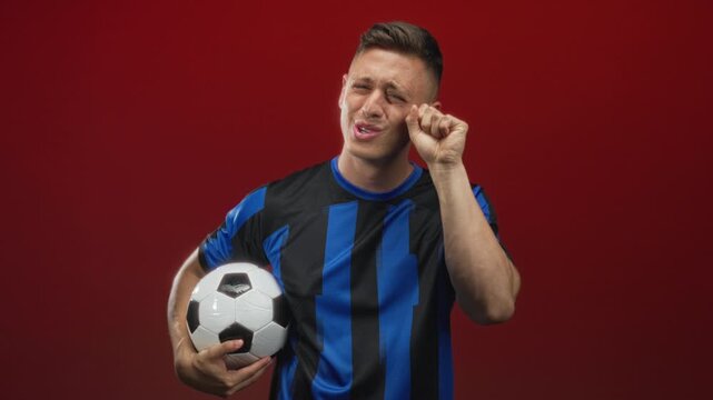 Man holds soccer ball and rubs eye with hand in studio red backdrop, striped blue jersey visible; sadness defeat.