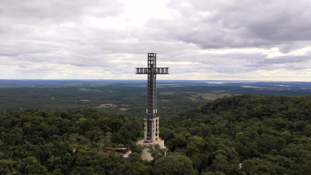 Monumental cross tower rises above Atlantic Forest landscape in Parque Tem&aacute;tico de la Cruz, Santa Ana, Misiones Province, Northeast Argentina, drone push ascent aerial shot beneath overcast sky