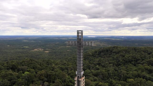 Monumental cross tower rises above Atlantic Forest landscape in Parque Tem&aacute;tico de la Cruz, Santa Ana, Misiones Province, Northeast Argentina, drone orbit establishing shot beneath subtropical clouds