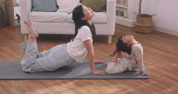 Cute Asian little girl doing yoga cobra pose on a mat next to her mother in the living room.