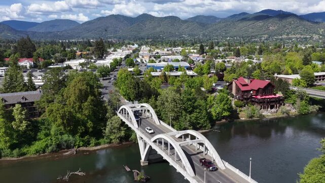 Grants Pass Oregon white arch bridge with truck crossing over Rogue River, red-roof historic building among dense green trees, low-rise downtown and mountain range backdrop. Aerial approaching shot.