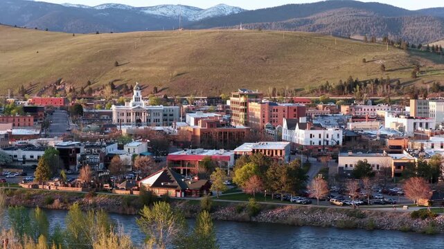 Missoula historic downtown with courthouse clock tower, brick buildings and Clark Fork River in foreground, bare spring trees lining riverbank and golden rolling hills. Montana, USA. Aerial wide shot