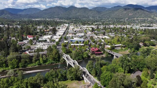 Grants Pass Oregon white arch bridge with truck crossing over Rogue River, red-roof historic building among dense green trees, low-rise downtown and forested mountain range backdrop. Aerial back wide