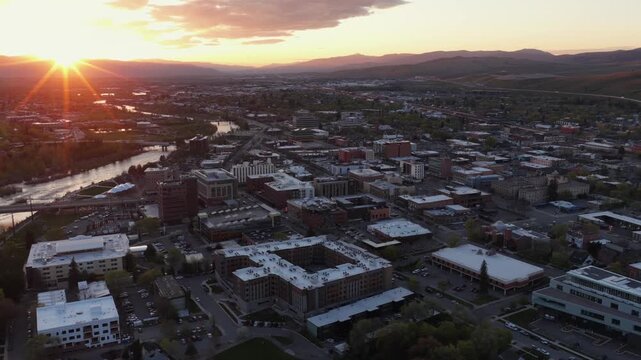 Missoula sprawling downtown and residential neighborhoods at golden sunset, Clark Fork River winding left, brick and modern buildings, suburban sprawl and rolling mountain range. Montana, USA. Aerial.