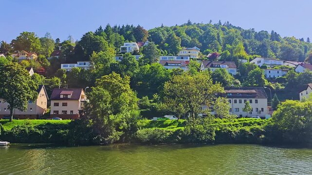 Heidelberg, Germany Neckar River view with a historic half timbered house, lush shoreline, green water, forested hills, warm sunlight, and a peaceful summer travel atmosphere by the water.
