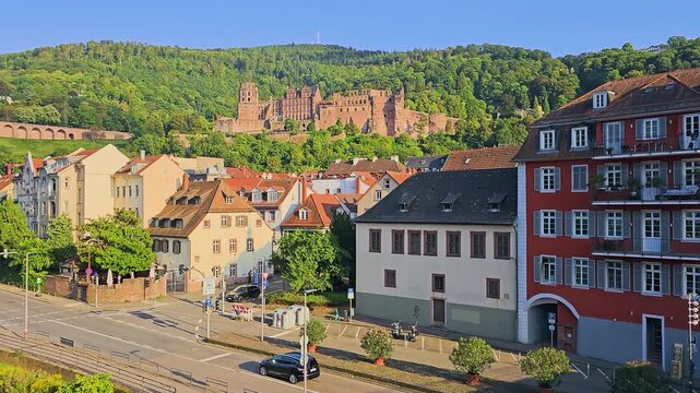 Historic Heidelberg, Germany skyline with castle ruins above colorful old town buildings, forested hill, red tile roofs, and clear blue sky in golden hour light for travel video.