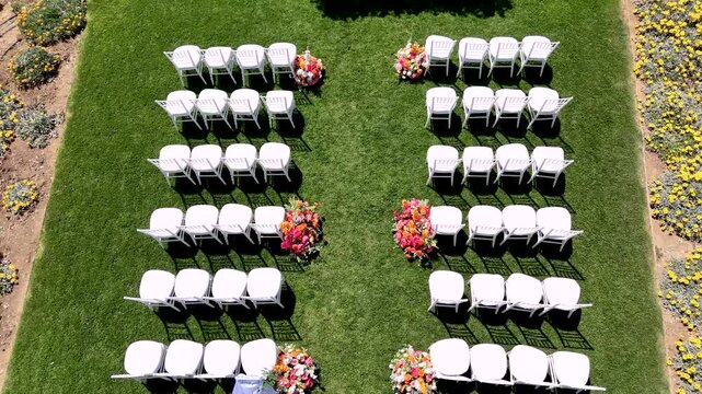 White chairs are set up in rows on green grass for a wedding event. Flowers are placed near the aisle. The setup shows a lovely garden location ready for the ceremony.