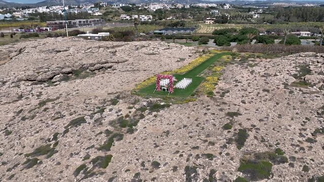 A couple stands in a decorated area filled with flowers on rocky ground. The beautiful landscape of Cyprus surrounds them. They share their special moment surrounded by nature under a clear sky.