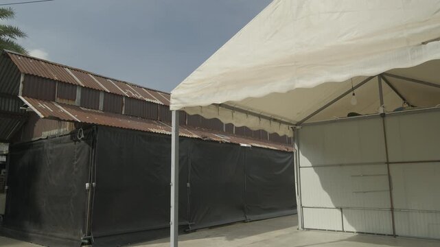 A wide shot of large white event tent structures set up next to a rustic, corrugated iron warehouse in an outdoor industrial area under a bright sky.