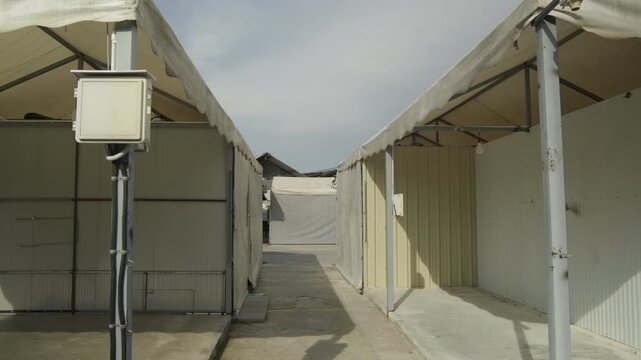 A cinematic shot moving through a narrow path between two large white event tents in an outdoor staging area, showing the structural frames and electrical equipment.