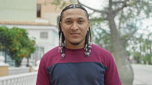 Man with beaded braids wearing maroon and navy sweatshirt smiles at camera on a street near houses and trees; friendly relaxed confidence.