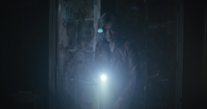 Scared young woman with flashlight entering dark room, propping door open and approaching small chest as storm rages outside