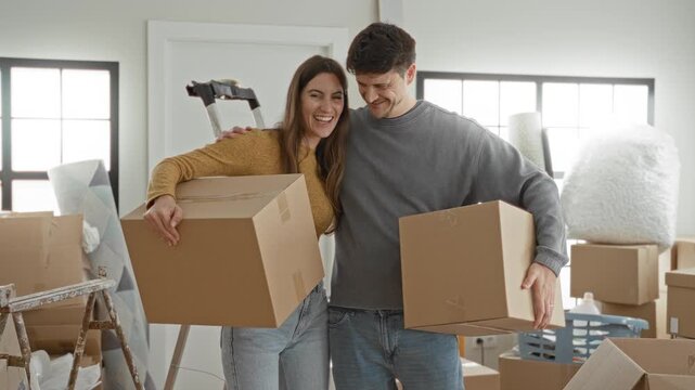 Young couple carrying cardboard boxes and smiling, woman leaning on man with arm around shoulder among packed furniture and ladder in a building move in scene; home joy.
