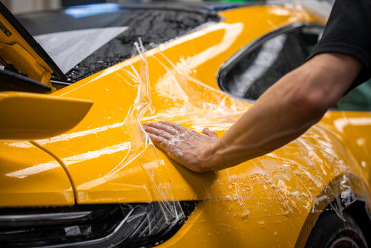 Inside a luxury car workshop, a technician smooths wet film on a yellow sports car rear fender, taillight and raised rear wing visible, under bright overhead lights.