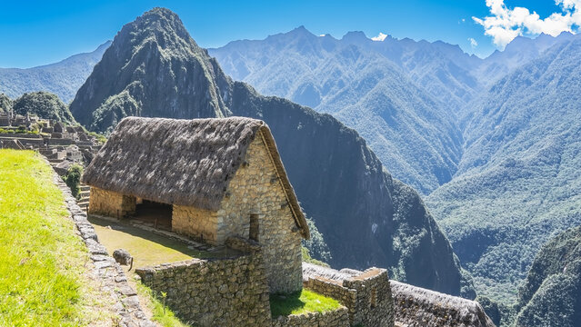 The famous Machu Picchu. On the mountainside, a masonry house with window openings, a thatched roof. Huayna Picchu, the majestic mountain range of the Andes against a blue sky and clouds. Peru. Cusco.