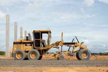Heavy Duty Grader on Construction Site: A yellow heavy-duty grader is actively leveling the ground on a construction site, with concrete pillars and a cloudy sky in the background. © Rassarin