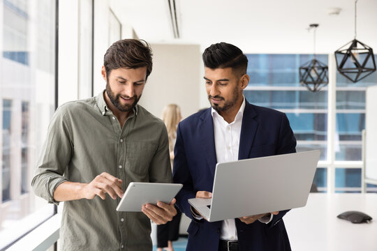 Two multinational male colleagues engaged in discussion in office boardroom, working with tablet and laptop, share ideas, reviewing or comparing data, take part in efficient teamwork, digital workflow
