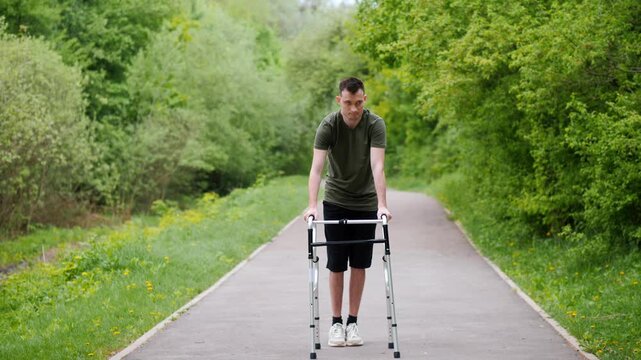 Young man with cerebral palsy learning to walk with walker on park path during rehabilitation process.