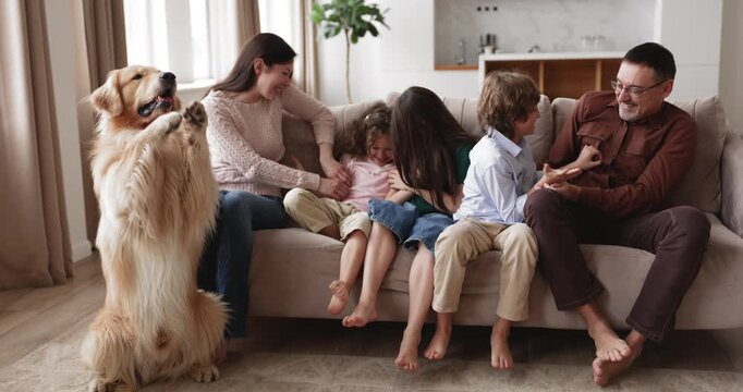 Happy large Caucasian family sit on couch, laughing, playing with kids. Parents tickling children, having fun, spending joyful leisure in living room with playful golden retriever dog at home. Bonding