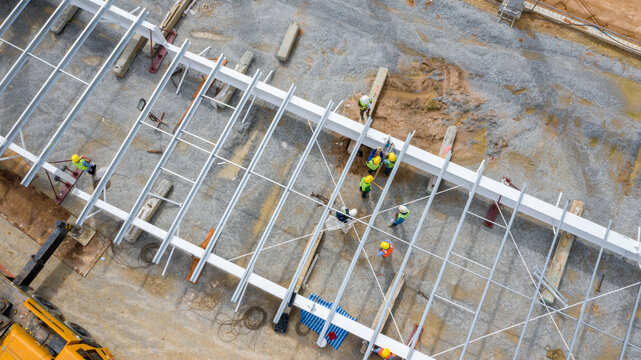 Industrial Construction Site: An aerial view of an industrial construction site shows workers in hard hats and high-visibility vests assembling a large metal structure.