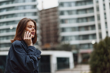 Fototapeta premium Young woman in a denim jacket talks on her smartphone outdoors in an urban setting, focused during a daytime call with a blurred city backdrop and cool tones.