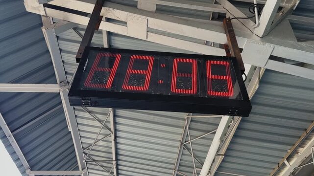 Dynamic camera orbiting around a digital LED clock display hanging inside an Indian railway station with metal roof structure and transportation infrastructure background.