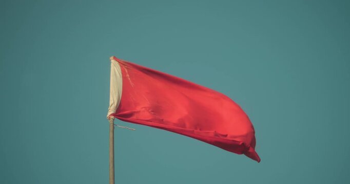 Red warning flag fluttering in strong coastal wind against blue sky. Beach safety symbol signaling dangerous swimming conditions and rough sea weather.
