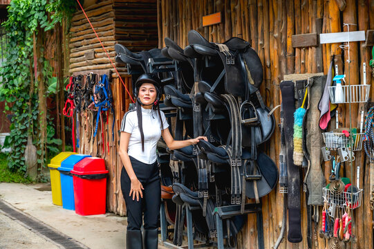 Female equestrian dressed in riding uniform  standing at stable tack room, selecting saddle from variety of horse saddles displayed on racks