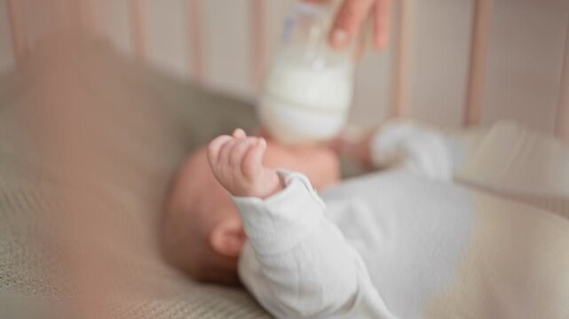 Baby boy drinking from bottle with mouth latched and hand raised while lying in crib in building; calm bonding.