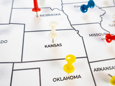 Close-up of a US map with an off-white pushpin on Kansas, a yellow pin on Oklahoma, a blue pin on Iowa, and red pins on Nebraska and Missouri. The white paper shows state borders and black labels.