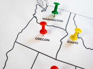 Close-up of a US map with a red pushpin on Oregon, a green pin on Washington, and a yellow pin on Idaho. The white paper shows state borders and names, shot with a shallow depth of field. © RODWORKS