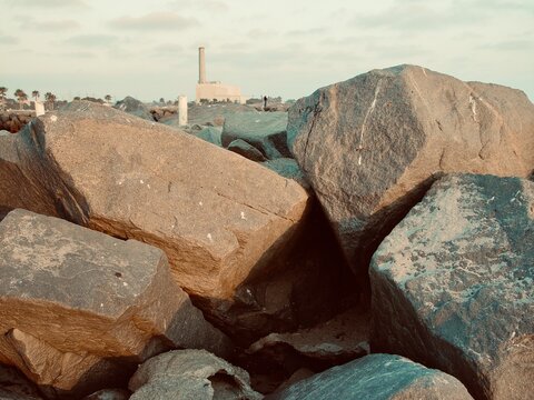 A unique structured landscape view of the old industrial Encina Power Plant building seen through a natural gap between two large riprap boulders in Carlsbad, California.