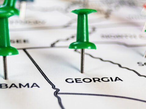 Macro shot of a green push pin on a paper map marking Georgia. Multiple surrounding green pins and clear state borders symbolize southern travel destination tracking, logistics, and road trips.