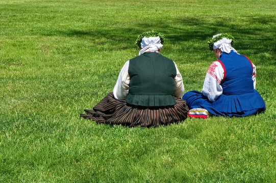  Two women in traditional Baltic folk costumes sitting on green grass. Midsummer festival celebration outdoors.