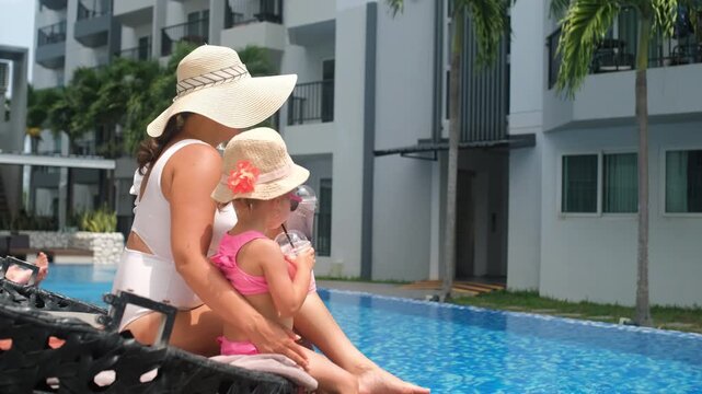 Side view of mother and little daughter both in straw hats and sunglasses sipping fruit cocktails together on a lounge chair beside the pool. Family holiday concept