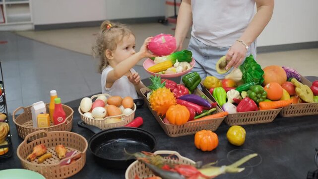 Mother and little daughter standing together at a colorful play kitchen counter selecting toy food items and plastic ingredients for pretend cooking. Family concept