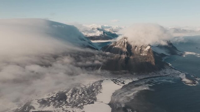 Aerial drone shot of Stokksnes in Iceland with snow covered mountains and coastline under a sunny sky with low clouds wrapping the peaks