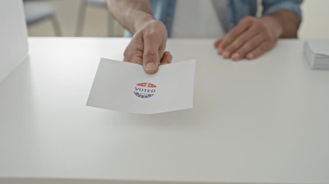 Man handing ballot with voted sticker over a white table inside a polling building, hand and paper clearly visible; civic pride.