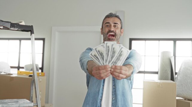 Man holds fan of dollars with both hands extended in building with moving boxes and ladder visible; celebration success.
