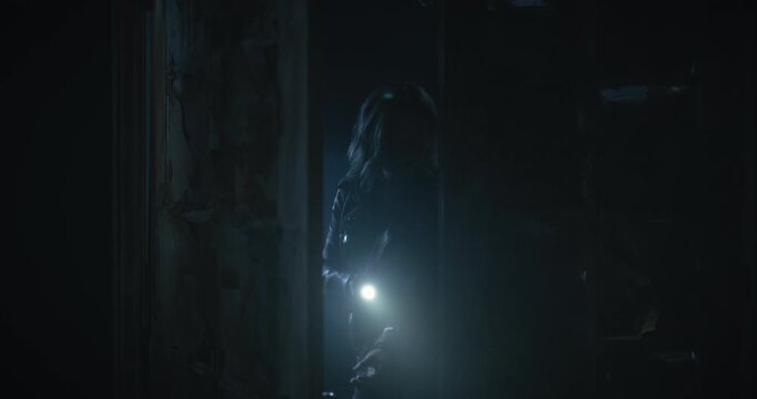Lone young woman entering dark, spooky room with flashlight as storm rages outside and she props door open behind her - medium shot
