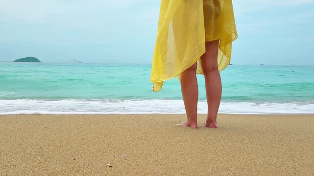 Woman in yellow pareo standing on wet sand with feet in shallow water, turquoise sea waves rolling ashore